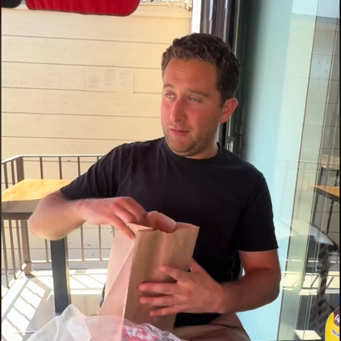 Person sitting at a table with a paper bag, a pickle on clear wrap, and a mustard bottle nearby