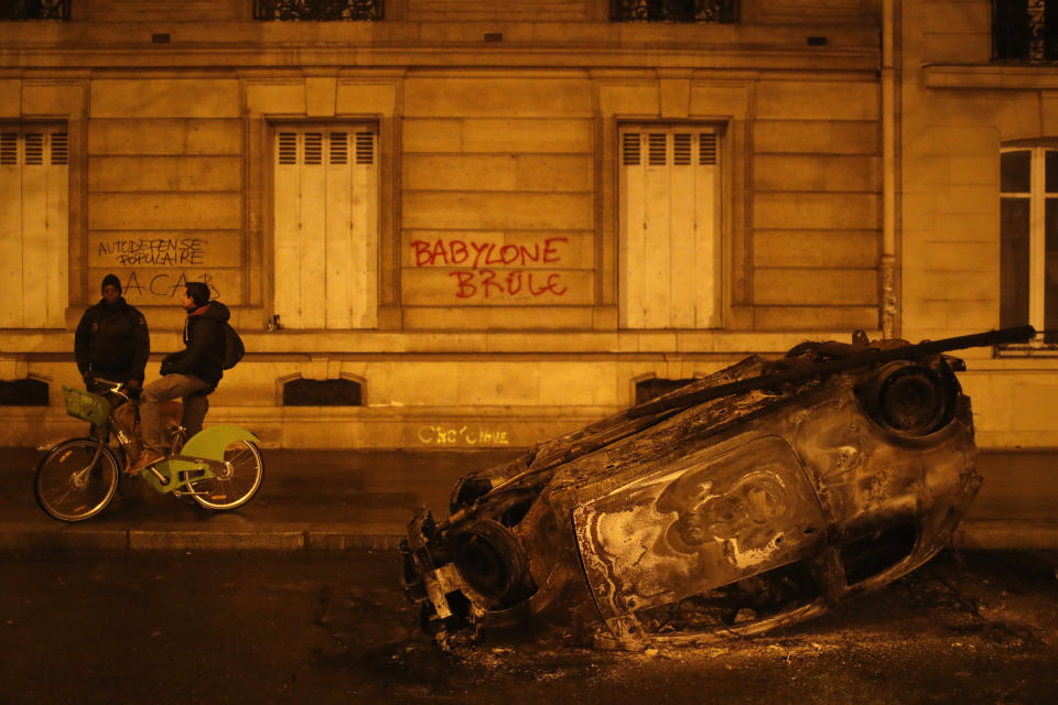 People talk next to a charred car with a graffiti reading "Babylon burns" on the wall after a demonstration Saturday, Dec.1, 2018 in Paris. A protest against rising taxes and the high cost of living turned into a riot Saturday in Paris as police fired tear gas and water cannon in street battles with activists wearing the fluorescent yellow vests of a new movement. (AP Photo/Thibault Camus)