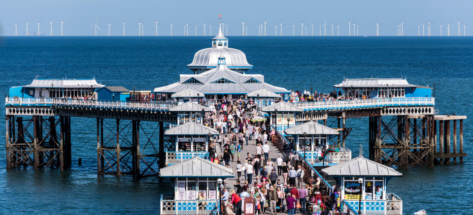 A view looking along Llandudno Pier on a busy summers day with the Wind Farm on the horizon beyond.