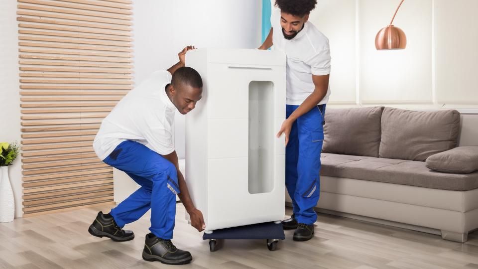 Two Young Male Movers Placing The White Cabinet On Wheel In The Living Room.