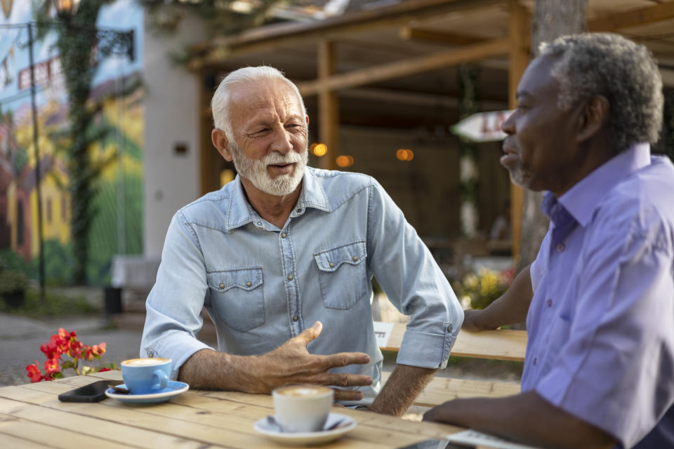 Two men, one in a light blue button-down shirt and the other in a purple short-sleeved shirt, chat in a street cafe with coffee cups on the table