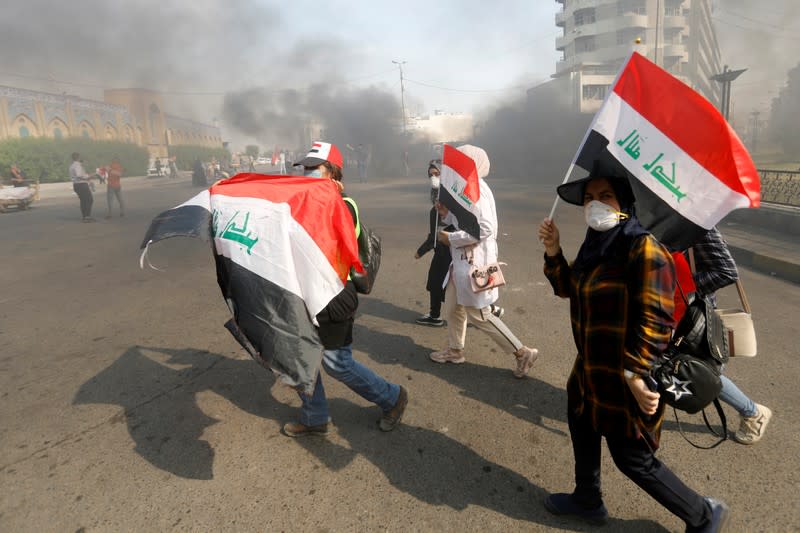 Iraqi demonstrators carry Iraqi flags as they walk at a cloud of smoke during ongoing anti-government protests, in Baghdad