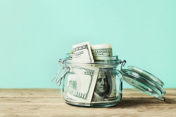 $100 bills stuffed in an open glass jar that's sitting on top of a wooden table