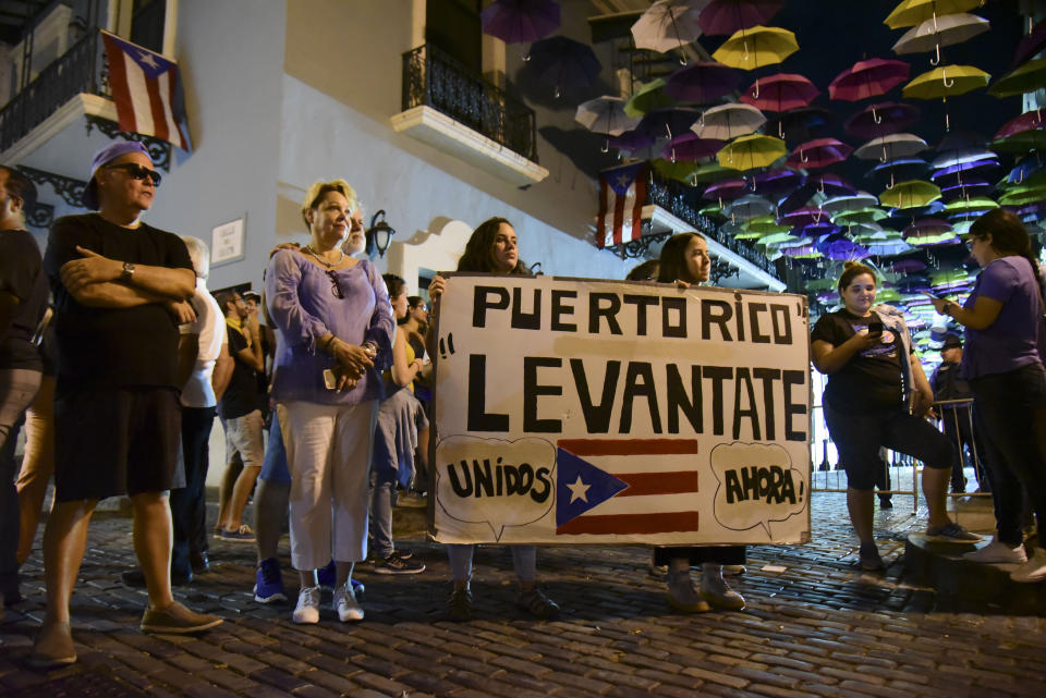 Dozens of citizens holding a sign that reads in Spanish "Puerto Rico rise up, unite now!" protest near the executive mansion denouncing a wave of arrests for corruption that has shaken the country and demanding the resignation of Gov. Ricardo Rosello, in San Juan, Puerto Rico, Thursday, July 11, 2019. Puerto Rico's former secretary of education and 5 other people have been arrested on charges of steering federal money to unqualified, politically connected contractors. U.S. Attorney for Puerto Rico Rosa Emilia Rodríguez said Gov. Rossello was not involved in the investigation. (AP Photo/Carlos Giusti)