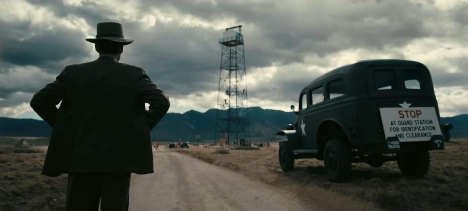 Cillian Murphy in a scene from "Oppenheimer" where J. Robert Oppenheimer is standing by the side of a dirt road next to a black car while gazing into the distance.