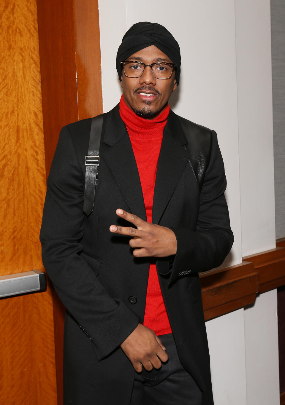 Man in black coat and red turtleneck posing with hand gesture