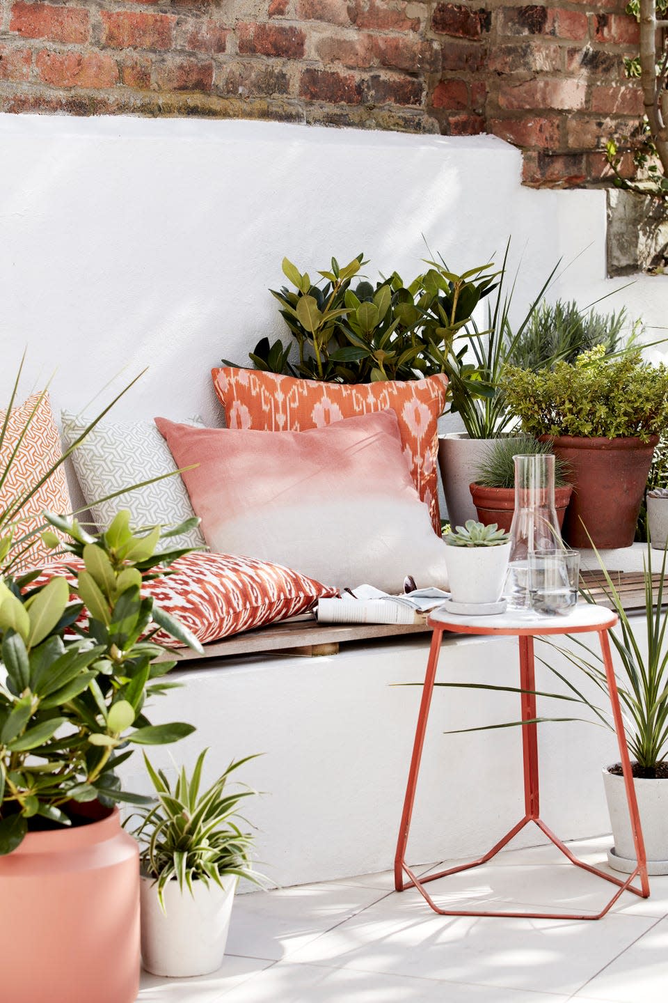 outside seating area, white bench with a wooden top covered in orange patterned cushions and a small table surrounded by pot plants instant oasis transform a shaded garden nook with piles of ikat print cushions in warming hues of burnt ochre and planters filled with lush foliage