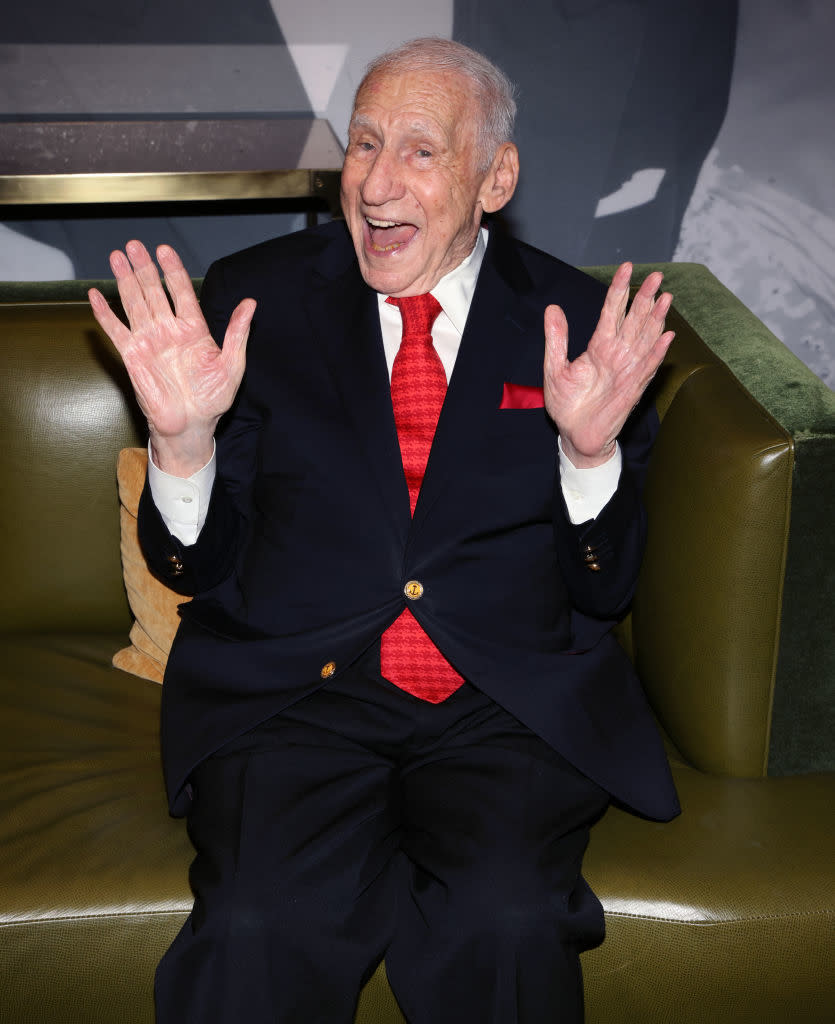 Close-up of Mel smiling and sitting on a sofa with his hands raised