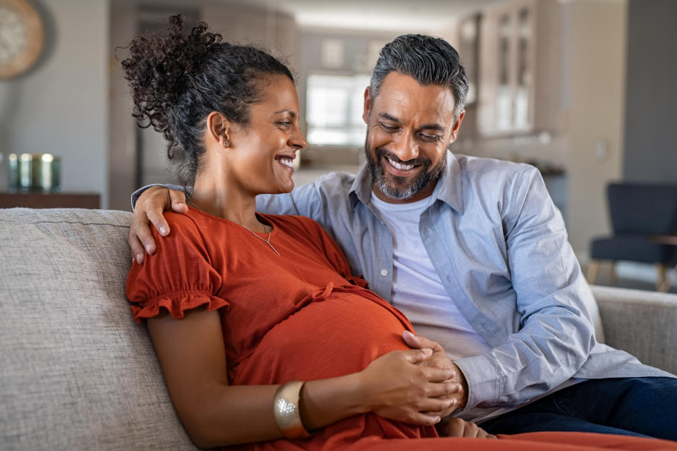 A pregnant woman and a man sit on a sofa as they smile and caress her belly