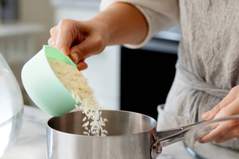 a woman's hands pouring a cup of uncooked white rice into a pot for cooking