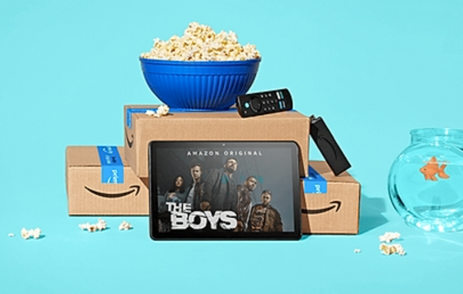 A blue bowl of popcorn sits on top of two cardboard boxes with the Amazon Prime logo, near a TV remote, a fish bowl, a tablet showing the film The Boys on a blue background.