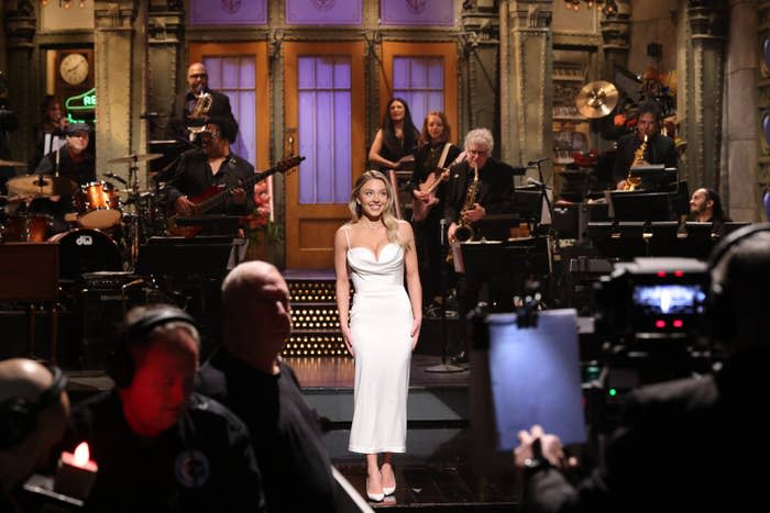 Woman in white dress onstage with musicians in the background, audience in the foreground, a cameraperson to the right