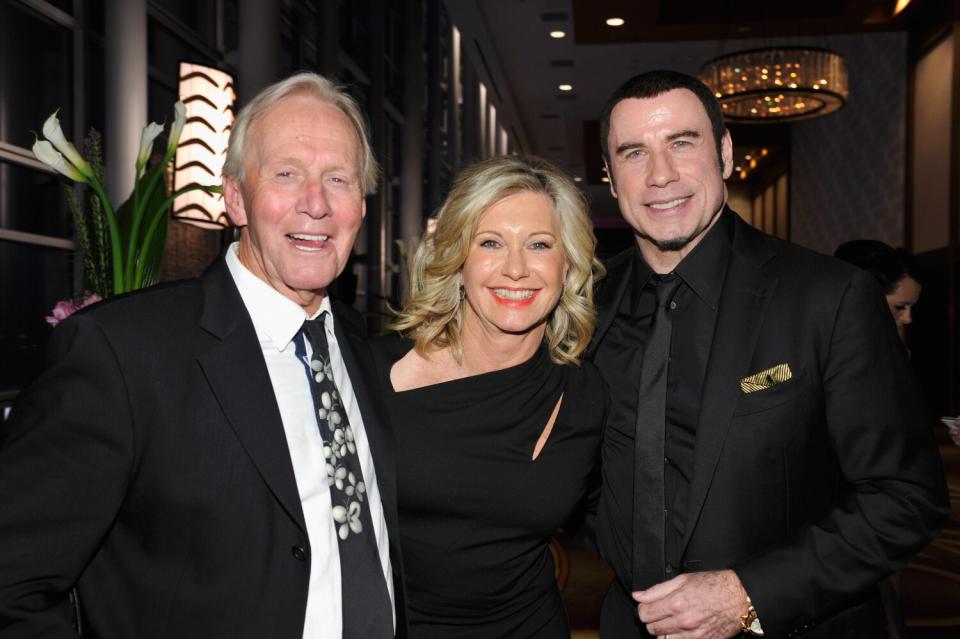 A smiling blond woman in a black dress stands between two men in suits at a gala event