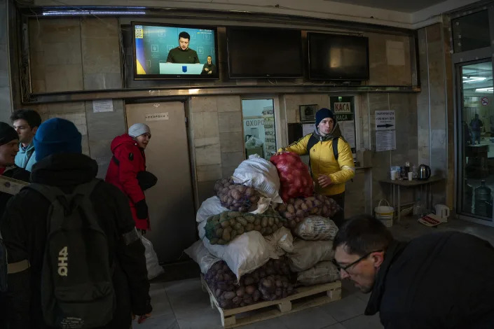 Volunteers in a small room process sacks of food donations while Ukrainian President Volodymyr Zelensky is seen on a television screen on a wall above them. 