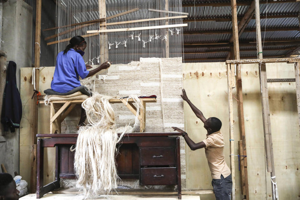 Manager of Texfad, John Baptist Okello, right,speaks to an employee as she weaves a carpet using banana fibre threads, at the Texfad factory in Sonde, Mukono District, Uganda, Sept. 20 2023. The decapitated banana plant is almost useless, an inconvenience to the farmer who must then uproot it and lay its dismembered parts as mulch. A Ugandan company is buying banana stems in a business that turns fiber into attractive handicrafts. The idea is innovative as well as sustainable in this East African country that’s literally a banana republic. (AP Photo/Hajarah Nalwadda)