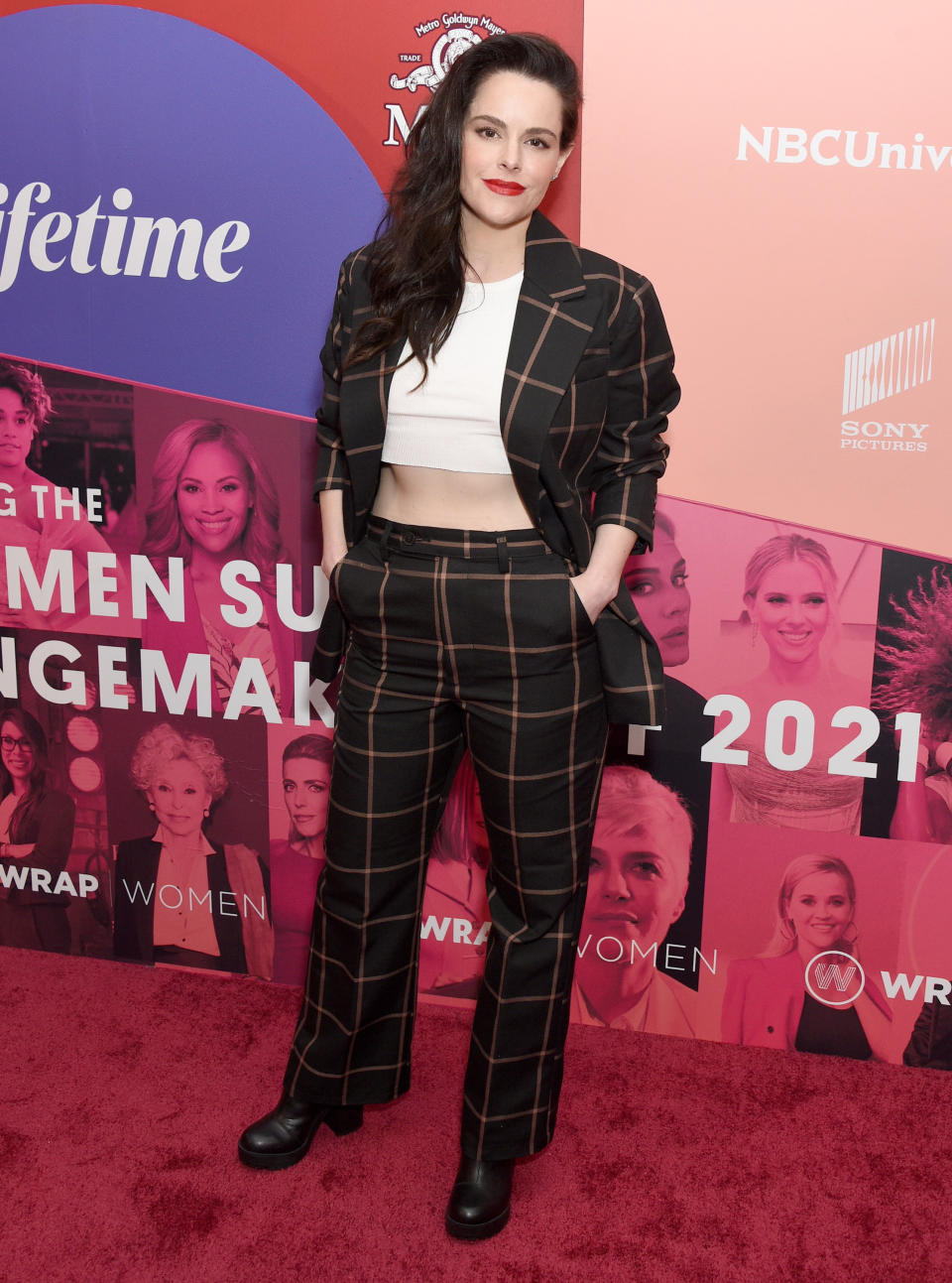 Emily Hampshire attends the WrapWomen's Power Women Summit & The Changemakers Of 2021. She wears a black suit with brown plaid pattern, with a white crop top underneath and chunky black boots.