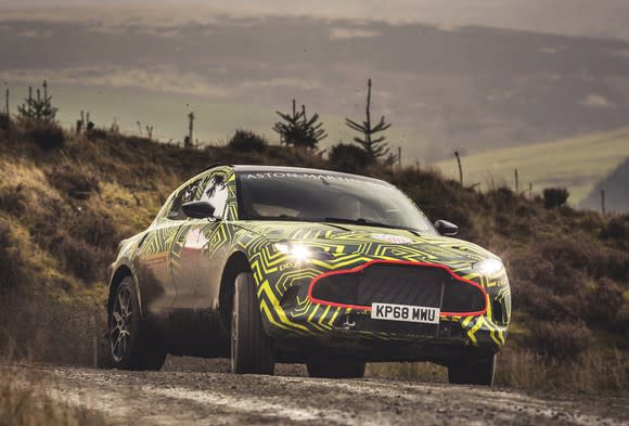 A prototype Aston Martin DBX, a high-performance luxury SUV, covered in yellow and black camouflage, in a rugged hilly setting.
