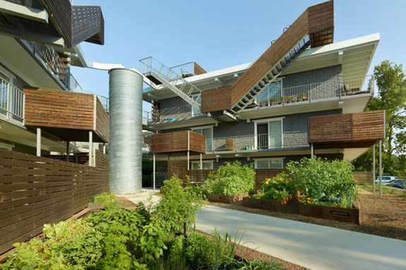 View of wedge courtyard at ECO Modern Flats with additive stairs, balconies and ground terraces. The rainwater harvesting cistern (silver cylinder) irrigates the community garden at the eastern entry to the site. The original buildings provided