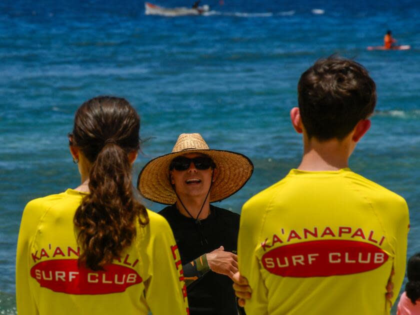 A person in a straw hat talks to two kids in Ka'anapali Surf Club T-shirts, the ocean behind them.