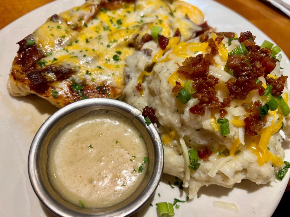 Chicken breast topped with melted cheese next to cup of white sauce and scoop of mashed potatoes topped with bacon bits, cheese, and chives at Outback Steakhouse