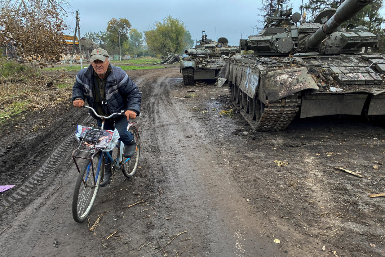 A resident rides a bicycle past abandoned Russian tanks on a muddy road.