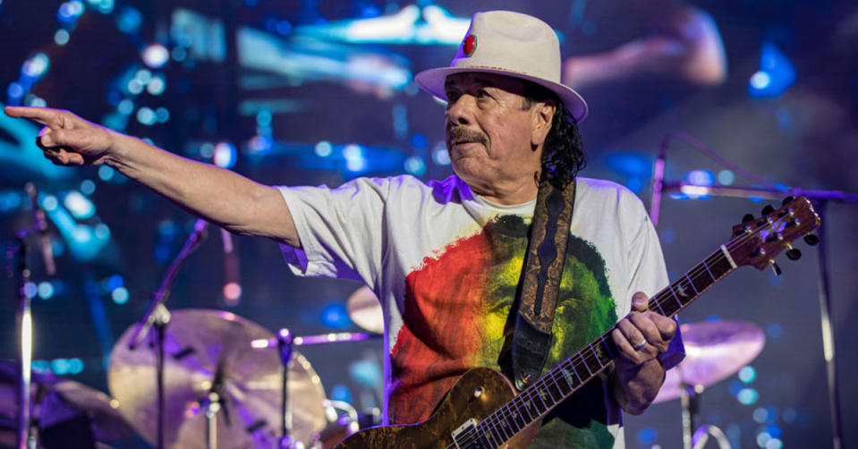 Carlos Santana holding a guitar, wearing a hat, on stage performing