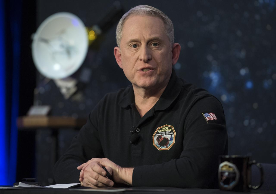 a man in a black shirt sitting at a table or desk with hands overlapped.