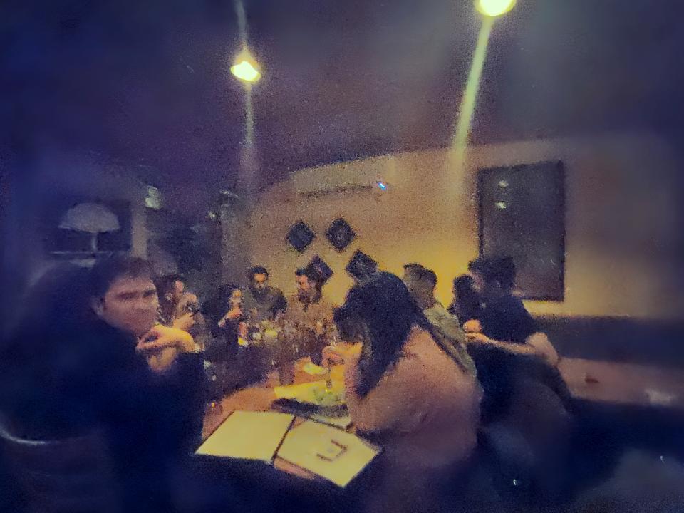 A sample image from the Humane AI Pin's 13-megapixel camera, showing a group of people sitting around a table in a dimly lit restaurant. One person is staring at the camera with his chin resting on the back of his hand. The photos is fuzzy and grainy.