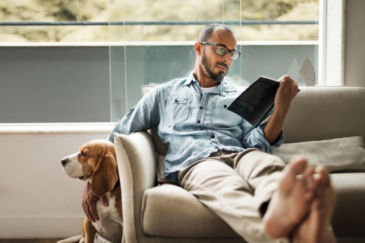 Bearded man comfortably sitting on a coach reading a book and holding his dog .