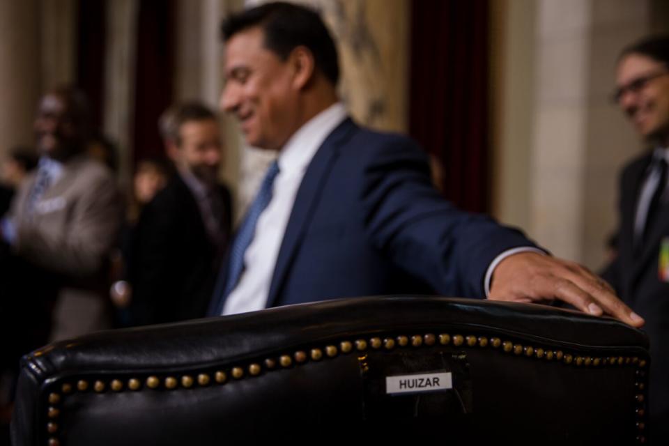 A man in a suit puts his hand on a chair with the label "Huizar"