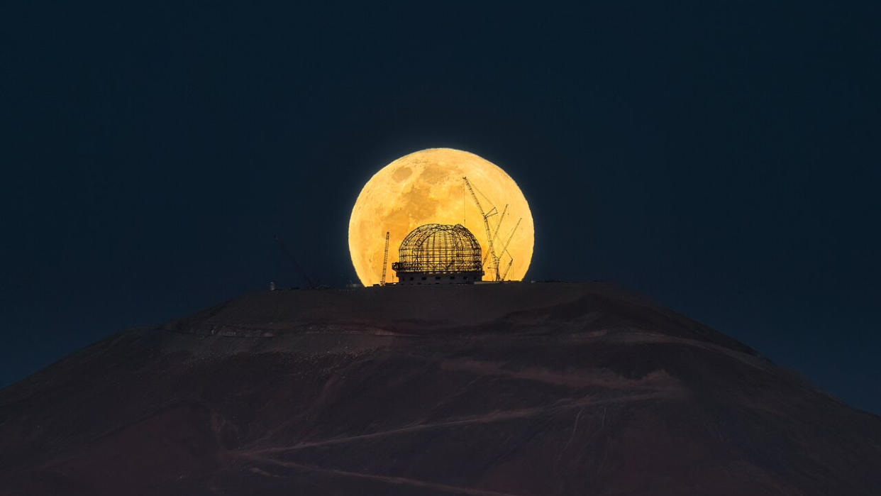  An orange moon rises from behind the top of a dark hill, shining through the scaffolding frame of a domed observatory. 