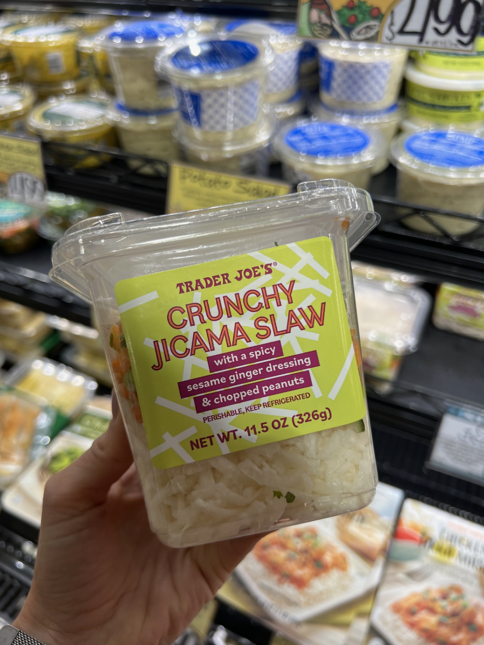 Hand holding a container of Trader Joe's Crunchy Jicama Slaw with a spicy sesame ginger dressing & chopped peanuts in a refrigerated grocery section