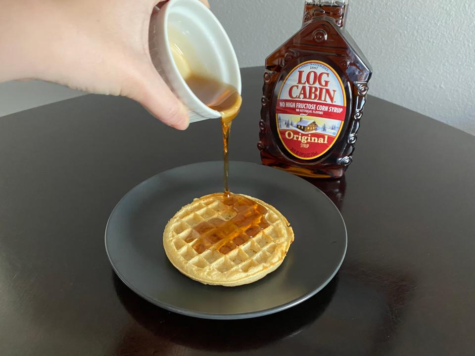 hand pouring white ramekin of log cabin syrup over a frozen waffle