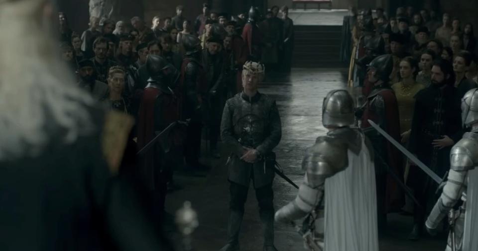 Viserys stands in front of Viserys in the Throne Room, with the Kingsguard between them and a crowd looking on