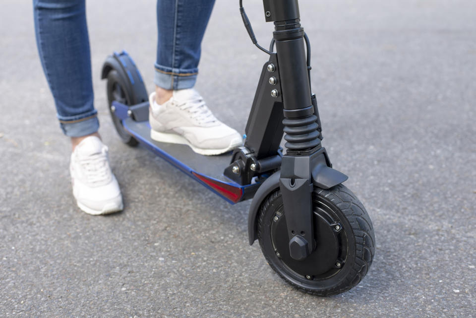 Person standing on an electric scooter, ready to ride, on a paved surface. The image is focused on the lower legs, scooter deck, and front wheel