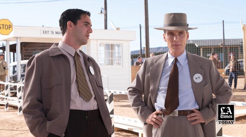 Two men stand outside talking, a fenced in compound behind them, in "Oppenheimer."