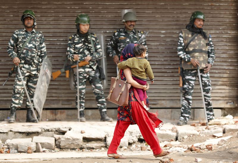 A woman carrying a child walks past security forces in a riot affected area after clashes erupted between people demonstrating for and against a new citizenship law in New Delhi