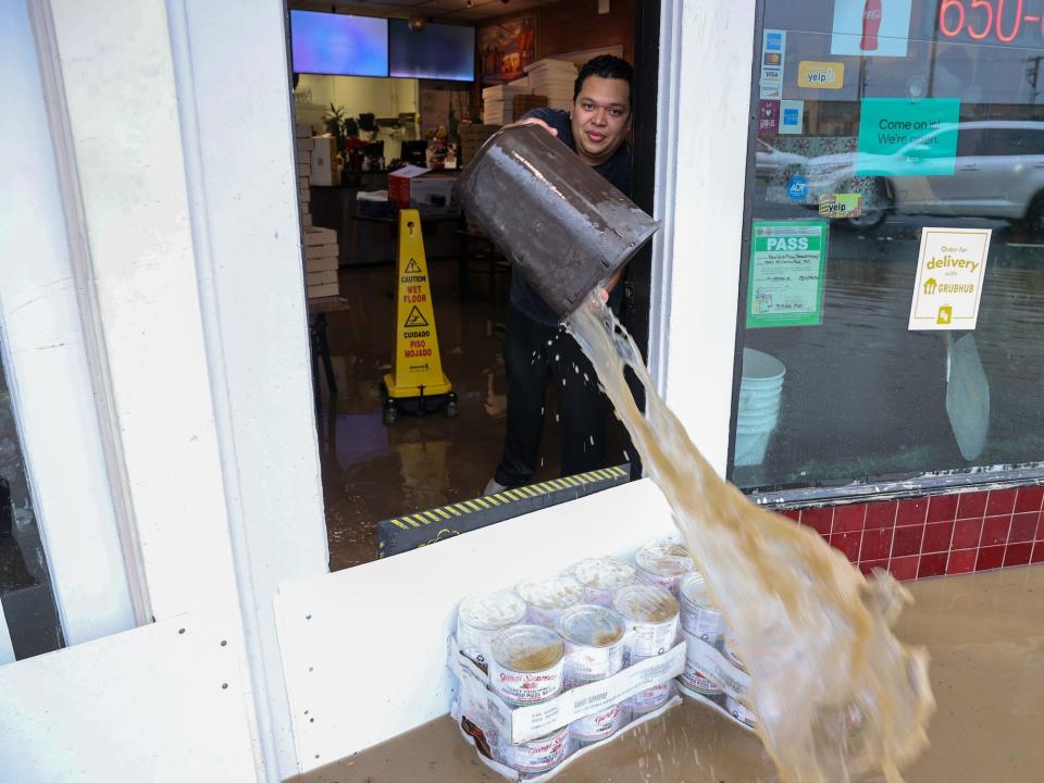 man dumps bucket of brown water onto flooded street through shop doorway
