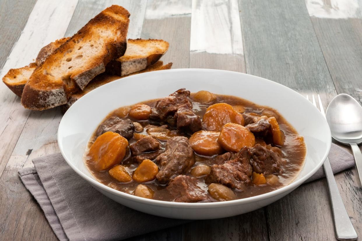 Ultimate slow cooker beef stew in a white bowl with three slices of toasted bread on a grey napkin with a fork and a spoon