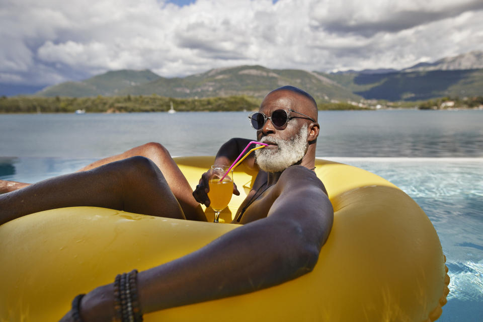 Man with a white beard and sunglasses relaxes on a yellow float in a pool while drinking an orange beverage through a colorful straw, with mountains in the background