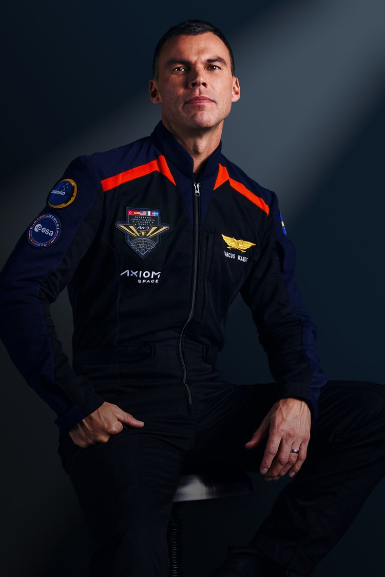 A man stands with his arms crossed, wearing a dark blue jumpsuit with wings on his shoulders with red accents.  He looks serious.