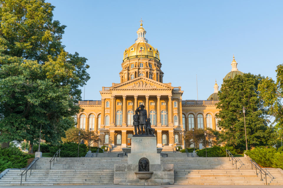 The image shows the Iowa State Capitol building with its grand architecture and dome, set against a clear sky. A statue stands prominently at the front