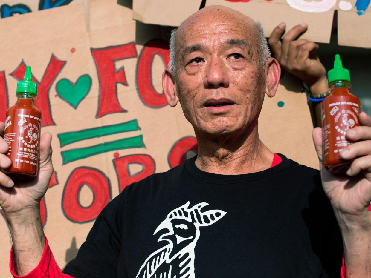 david tran, creator of sriracha, holds up two bottles of sriracha in each hand in front of a protestor's sign