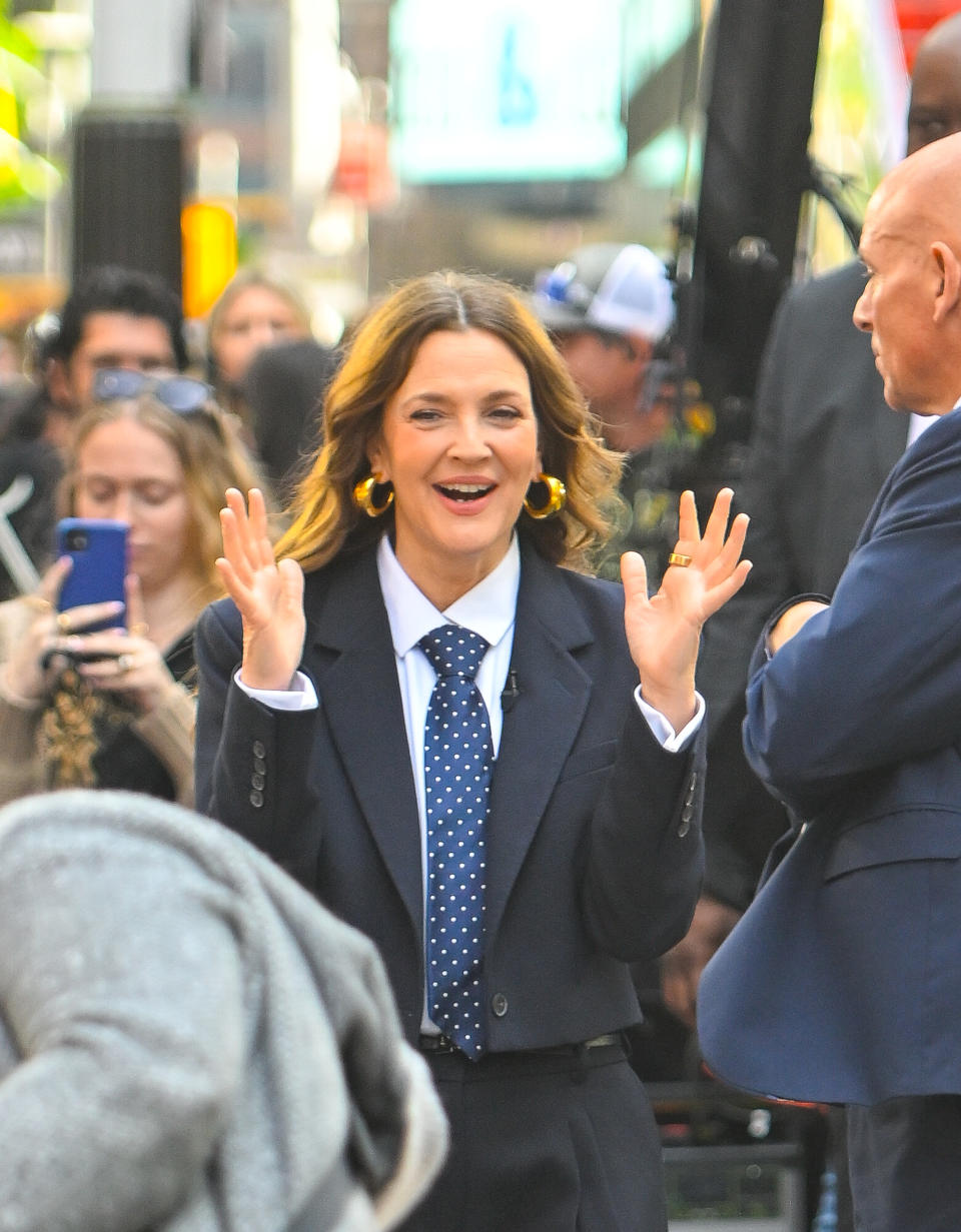 Drew Barrymore wearing a polka dot tie and blazer, smiling and gesturing with open hands while on a busy city street. People and cameras are visible in the background