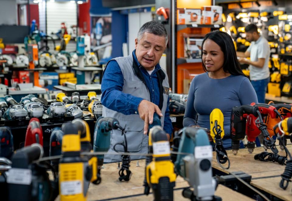 A-hardware-store-employee-talks-to-a-woman-about-the-power-drill-options-on-a-table.