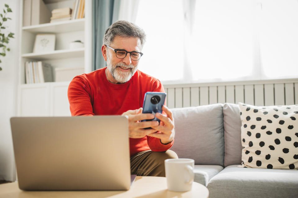 A man with glasses and beard, wearing a casual long-sleeve shirt, smiles while looking at his phone. He is sitting in front of a laptop on a table