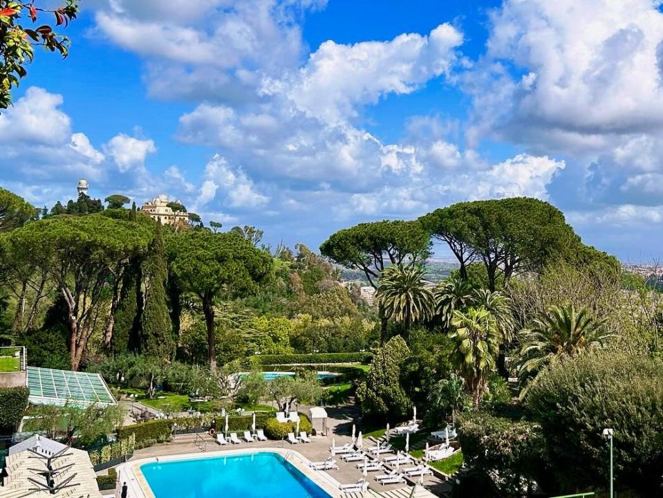 A bright-blue pool surrounded by white lounge chairs and lush green trees in foreground. There are bright-blue skies and clouds