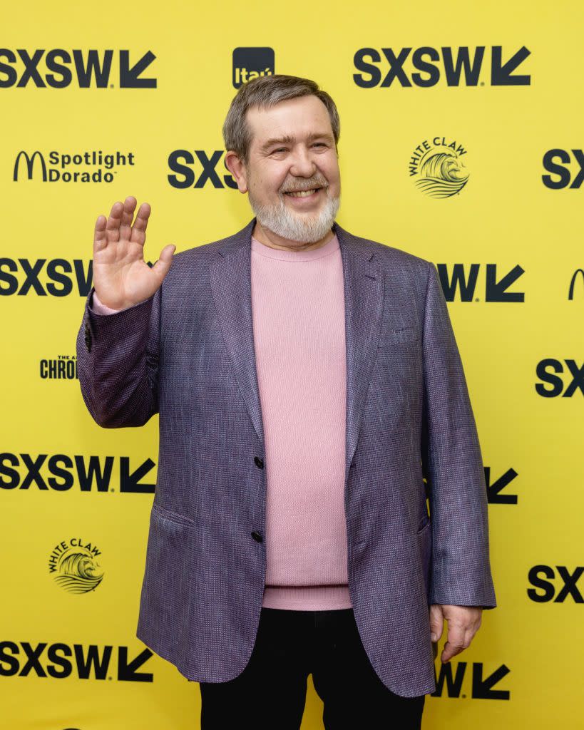 alexey pajitnov, wearing a gray suit jacket and pink shirt, stands in front of a yellow wall with several logos on it, smiling and waiving his right hand