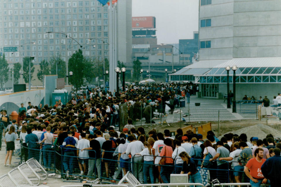 Large crowd of people forming a long line outside a building, likely waiting for an event or entry. Urban environment with buildings and construction visible