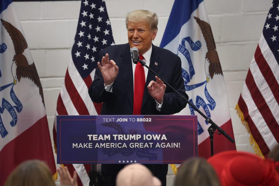 donald trump, wearing a black suit and red tie, standing at a podium and speaking to a crowd from a microphone, while standing in front of several flags